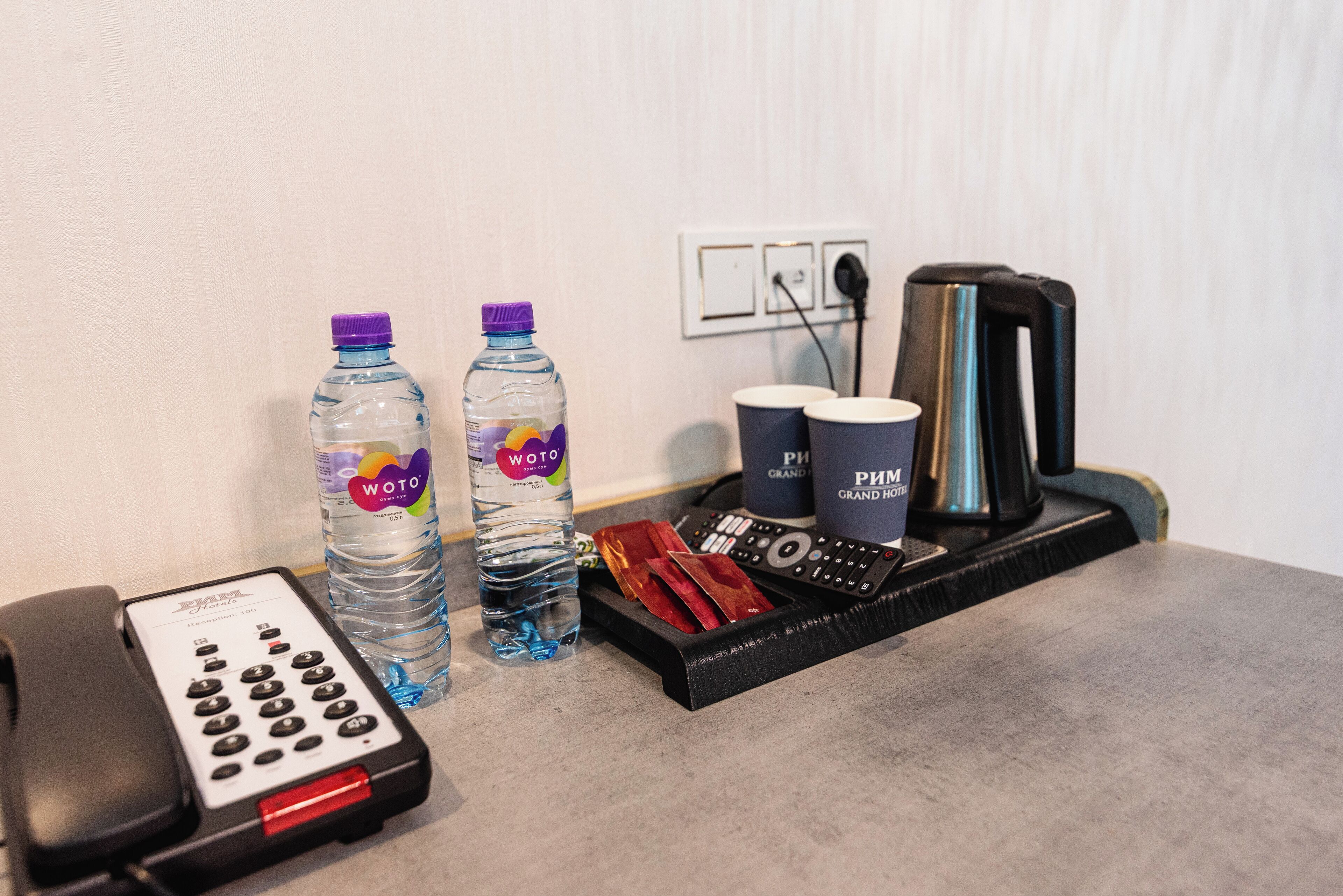 Escritorio de una habitación de hotel con un teléfono, dos botellas de agua, una bandeja con una cafetera, tazas y un control remoto.