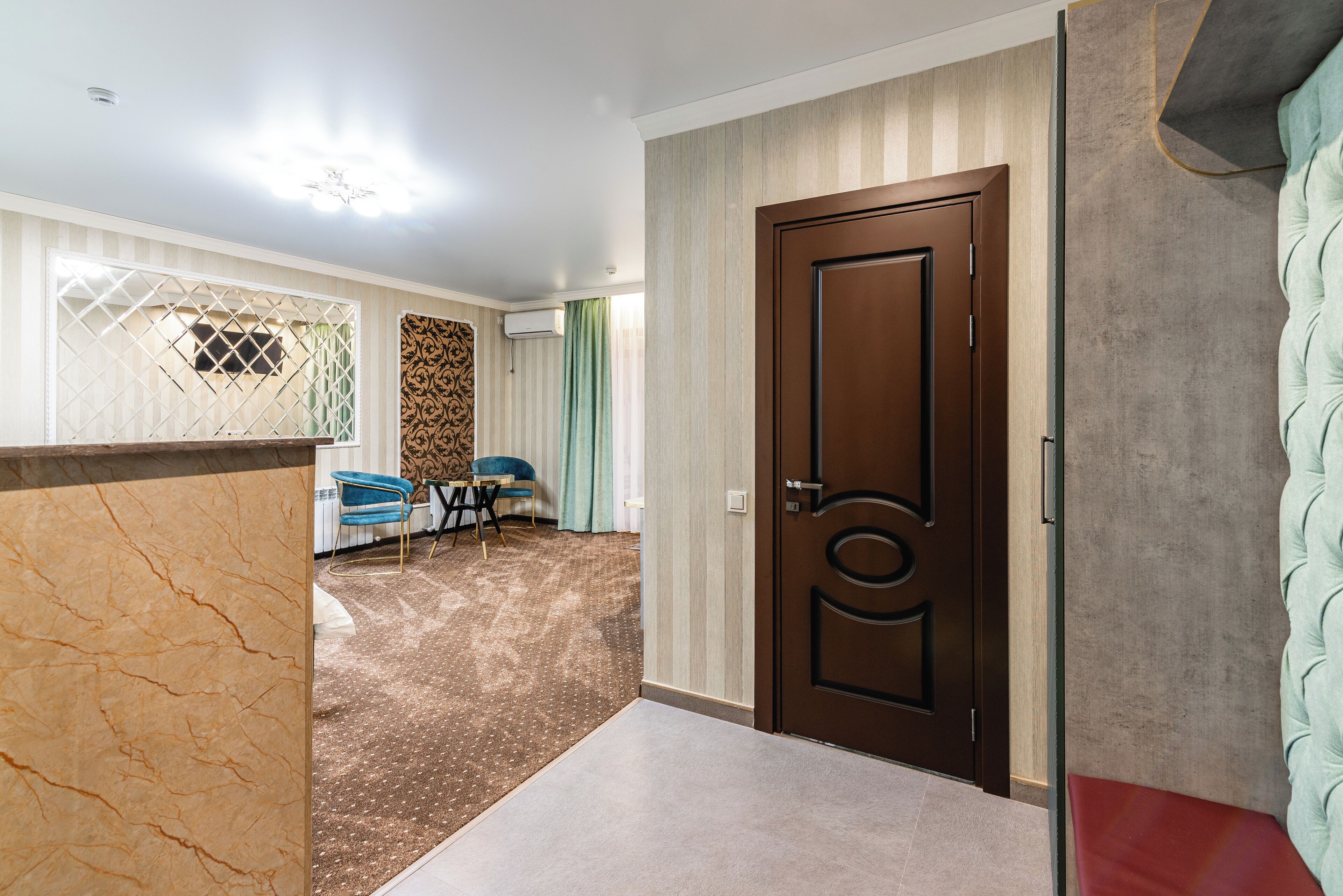 A hotel room with a wooden door, a marble accent wall, a patterned carpet, a blue chair, and a bed with a green headboard.