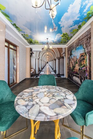 A hotel corridor with a mural, green upholstered chairs, a round table with a stone pattern, and a chandelier.