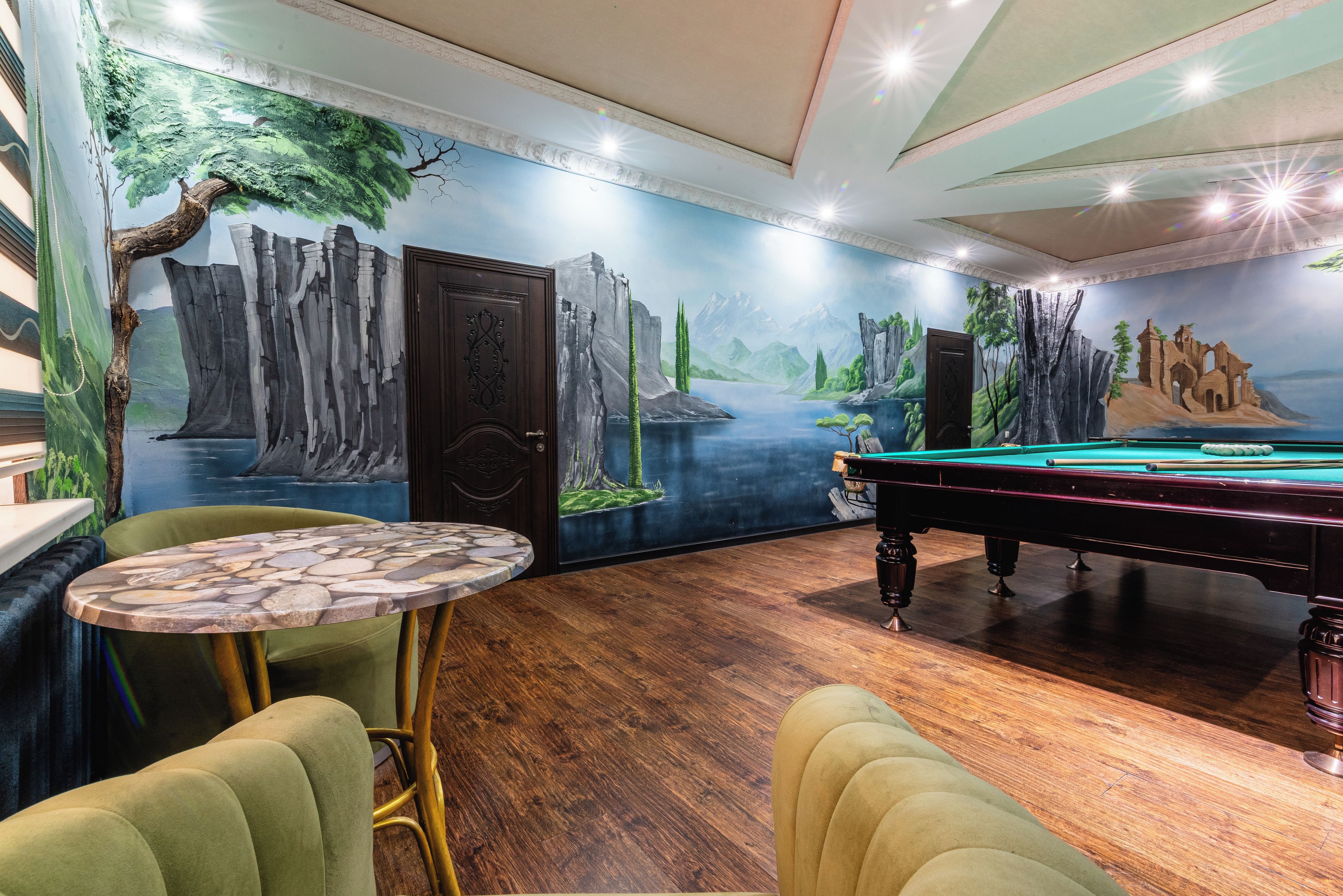 A room with a pool table, a green cushioned chair, a small round table with a stone pattern, and a mural depicting a forest scene.