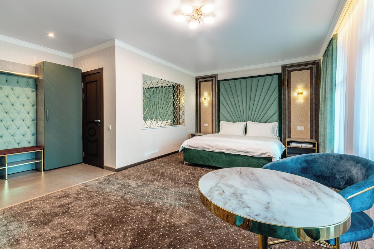 Una habitación de hotel con una cama, una mesa redonda de mármol, un sillón azul, un banco y un cabecero tapizado en verde.