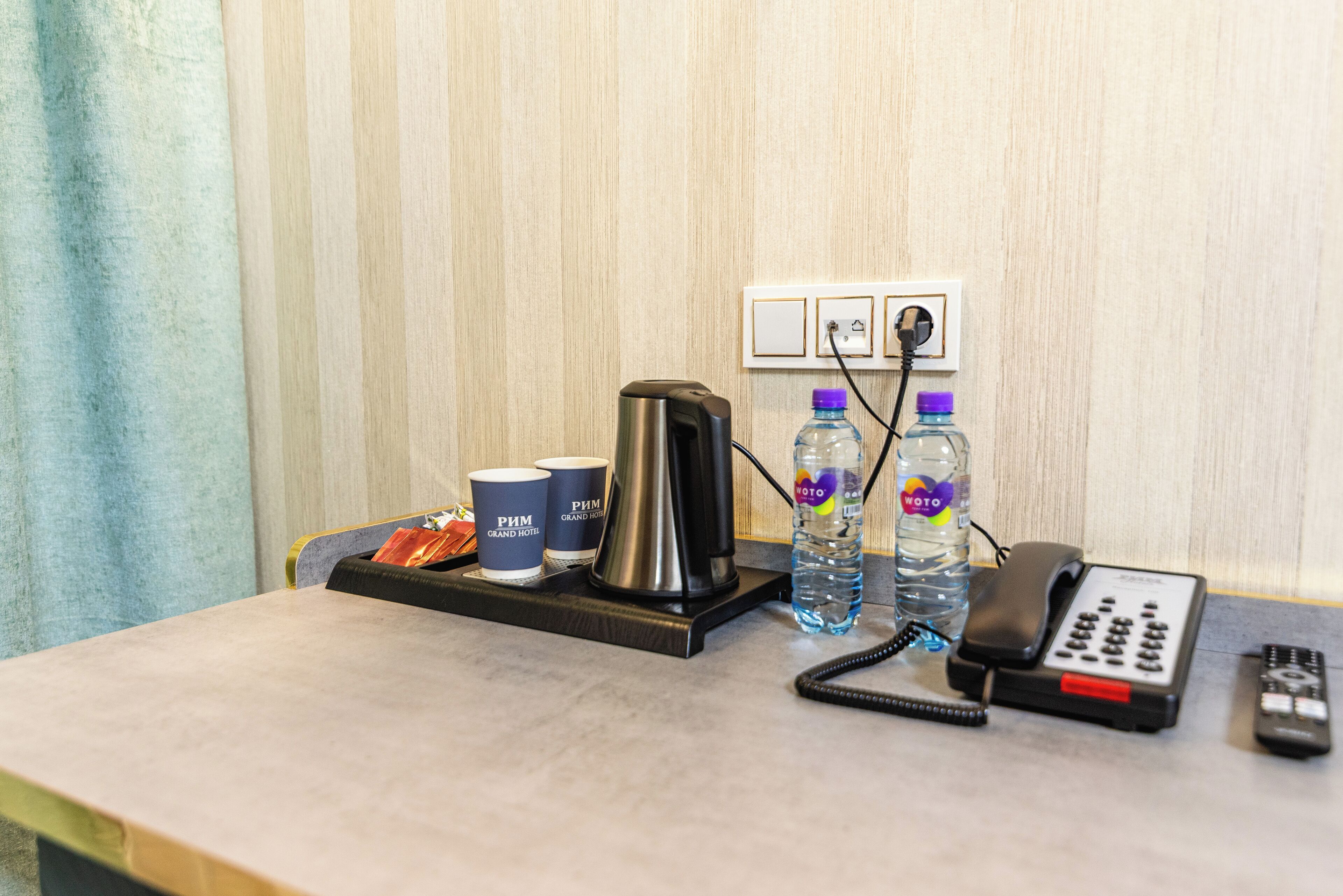 A hotel desk with a coffee maker, two cups, two water bottles, a phone, and a remote control.