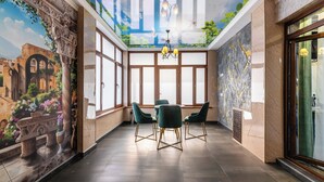 Une pièce spacieuse avec un plafond haut, de grandes fenêtres et une fresque représentant un jardin. On y trouve trois chaises vertes et une table ronde au centre.