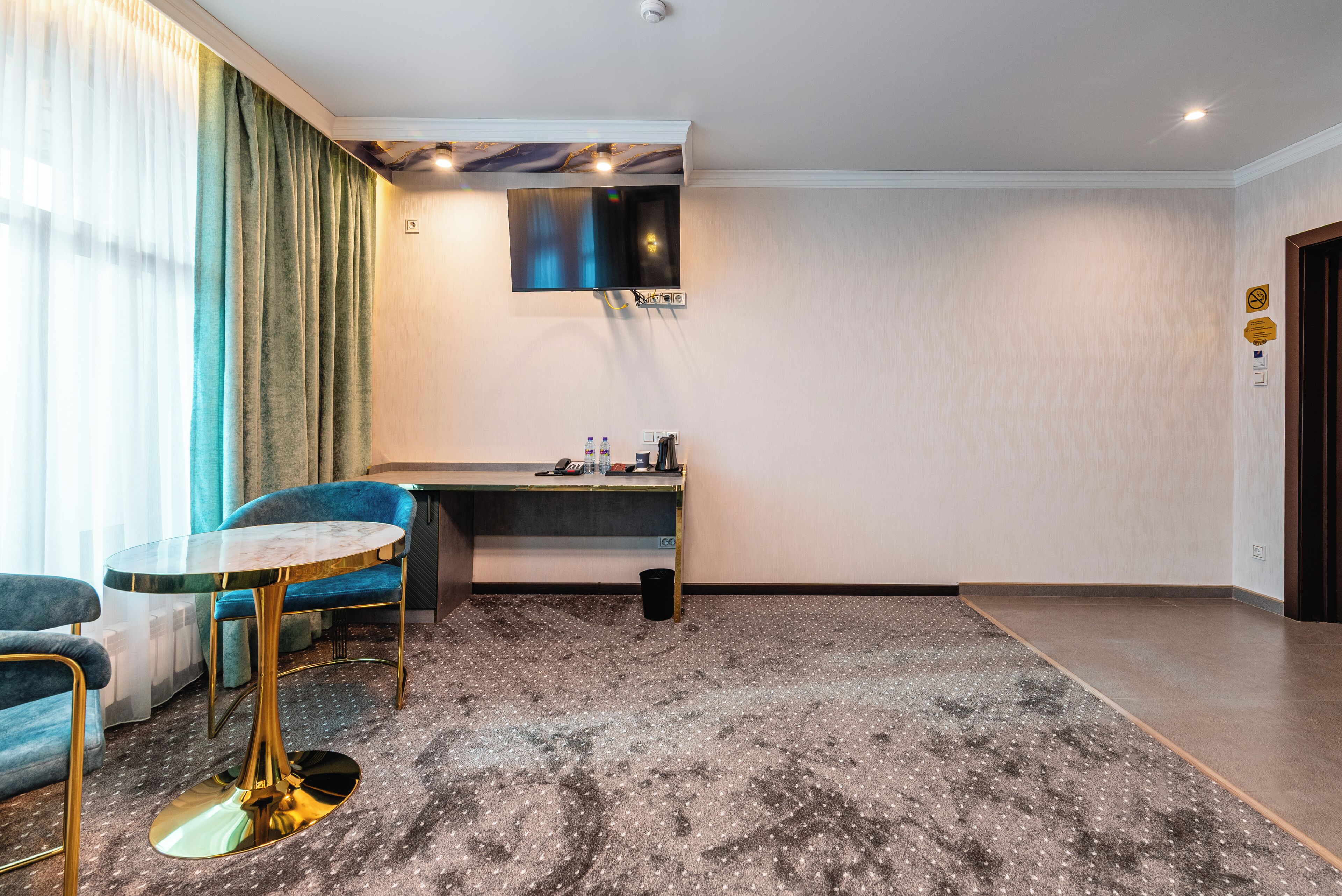 Habitación de hotel con una mesa redonda, dos sillones azules, un escritorio y un televisor de pantalla plana montado en la pared.