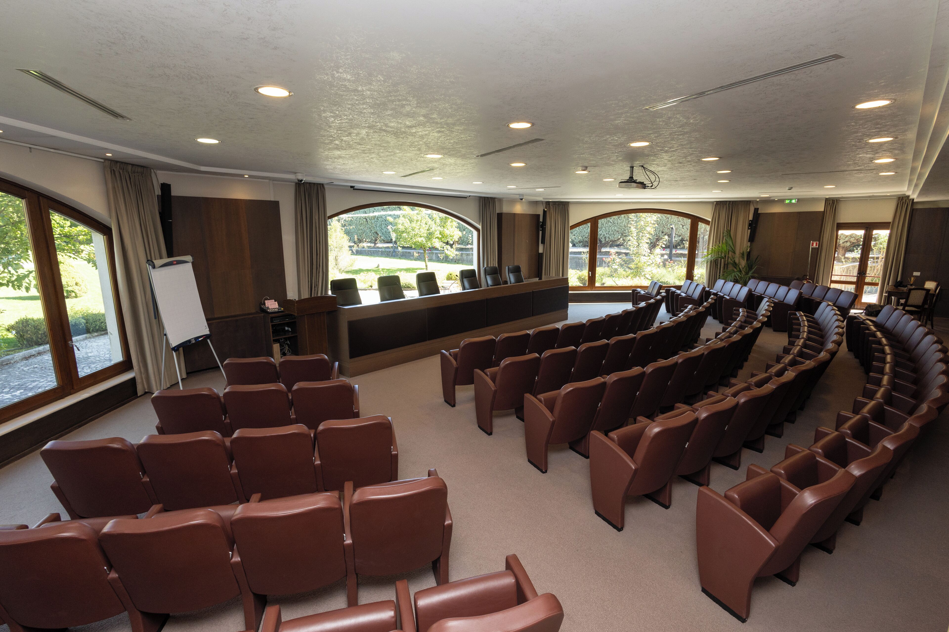 A conference room with rows of brown chairs, a whiteboard, and large windows offering a view of greenery.
