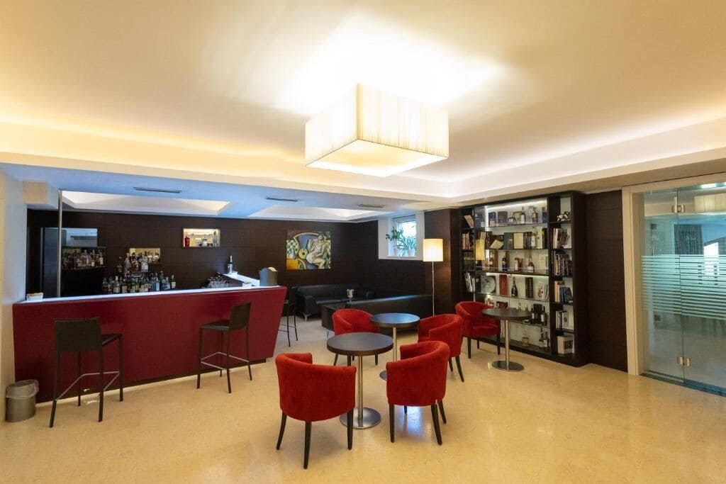 Uma área de bar moderna com balcão vermelho, cadeiras vermelhas e uma mesa redonda. Há uma estante com vários objetos à direita e uma pintura na parede atrás do balcão.