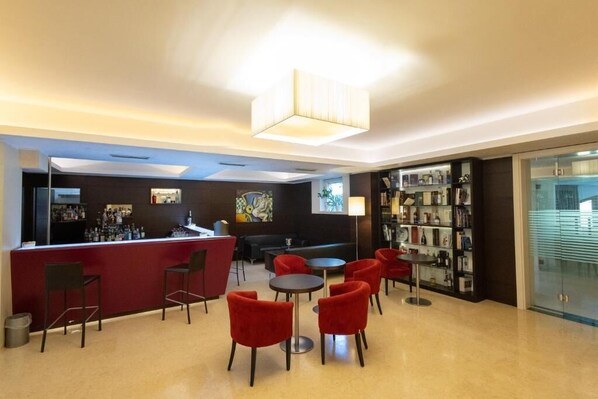 A modern bar area with a red counter, red chairs, and a round table. There is a bookshelf with various items on the right and a painting on the wall behind the bar. - Hotel La Dimora del Baco (L'Aquila)