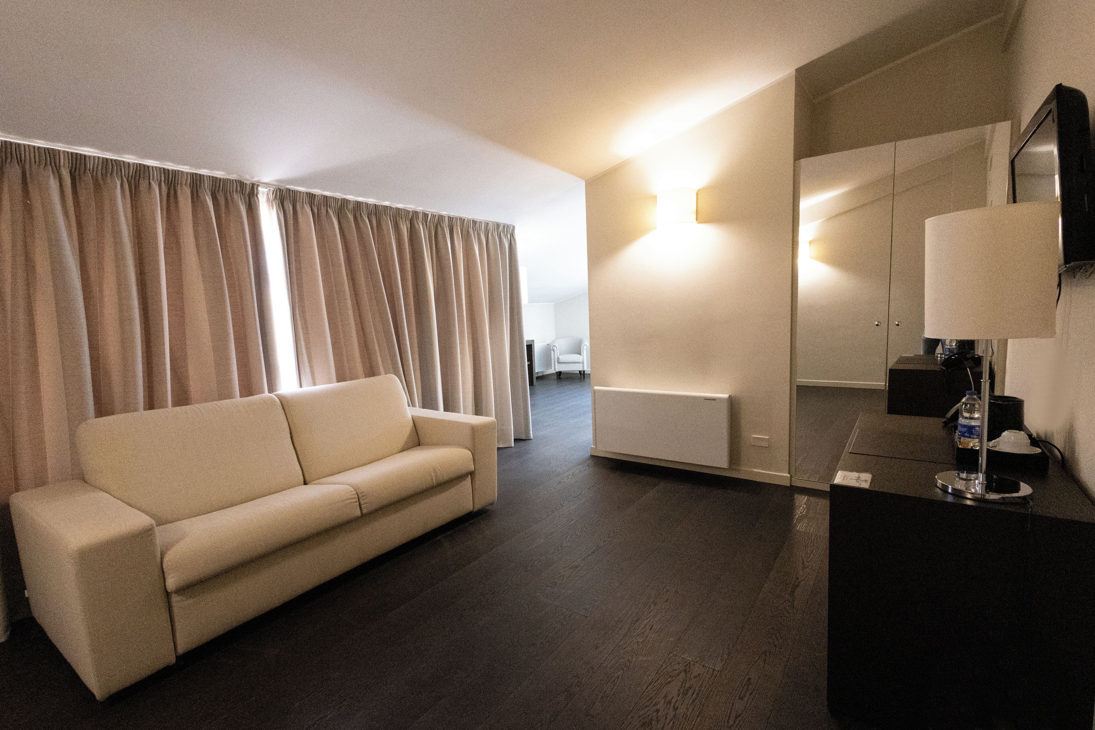 Una habitación de hotel moderna con un sofá color beige, piso de madera oscura, un espejo grande y una lámpara fijada a la pared.