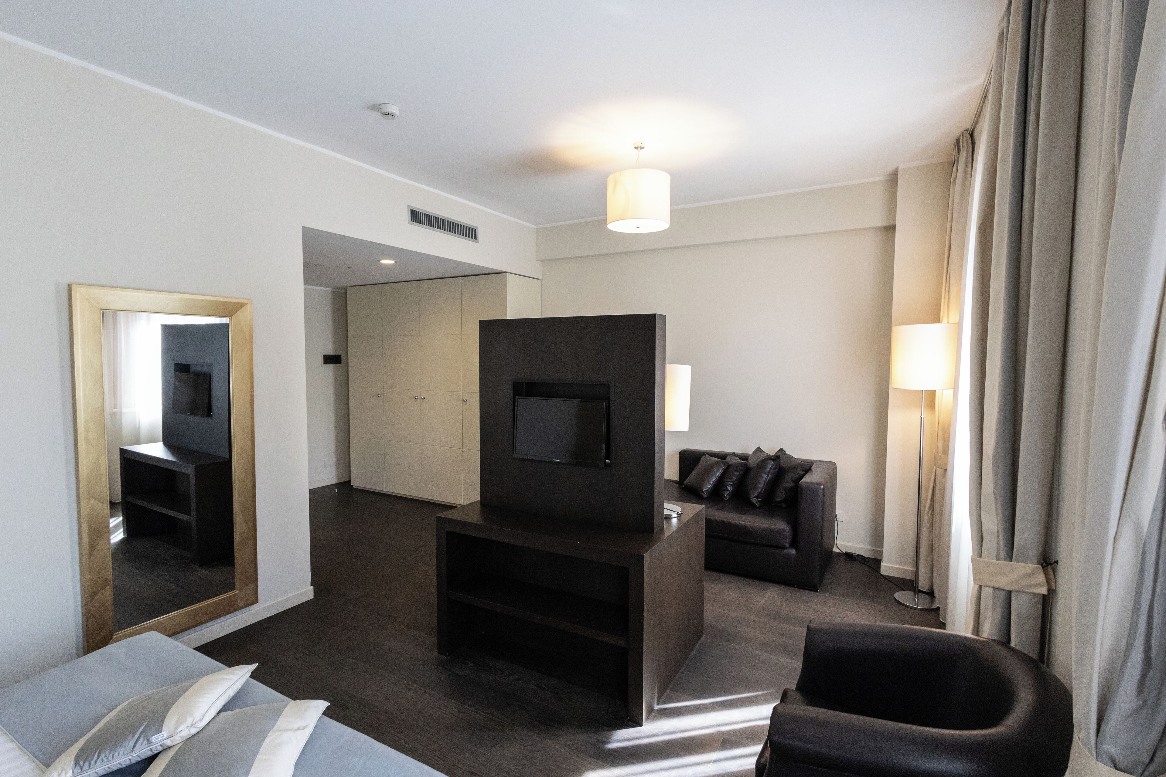 A modern hotel room with a flat-screen TV, a sofa, a chair, a large mirror, and a floor lamp.