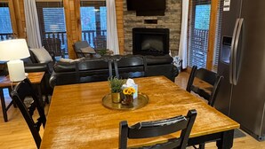 Une table à manger en bois avec des chaises, une cheminée, un téléviseur fixé au mur et un réfrigérateur.