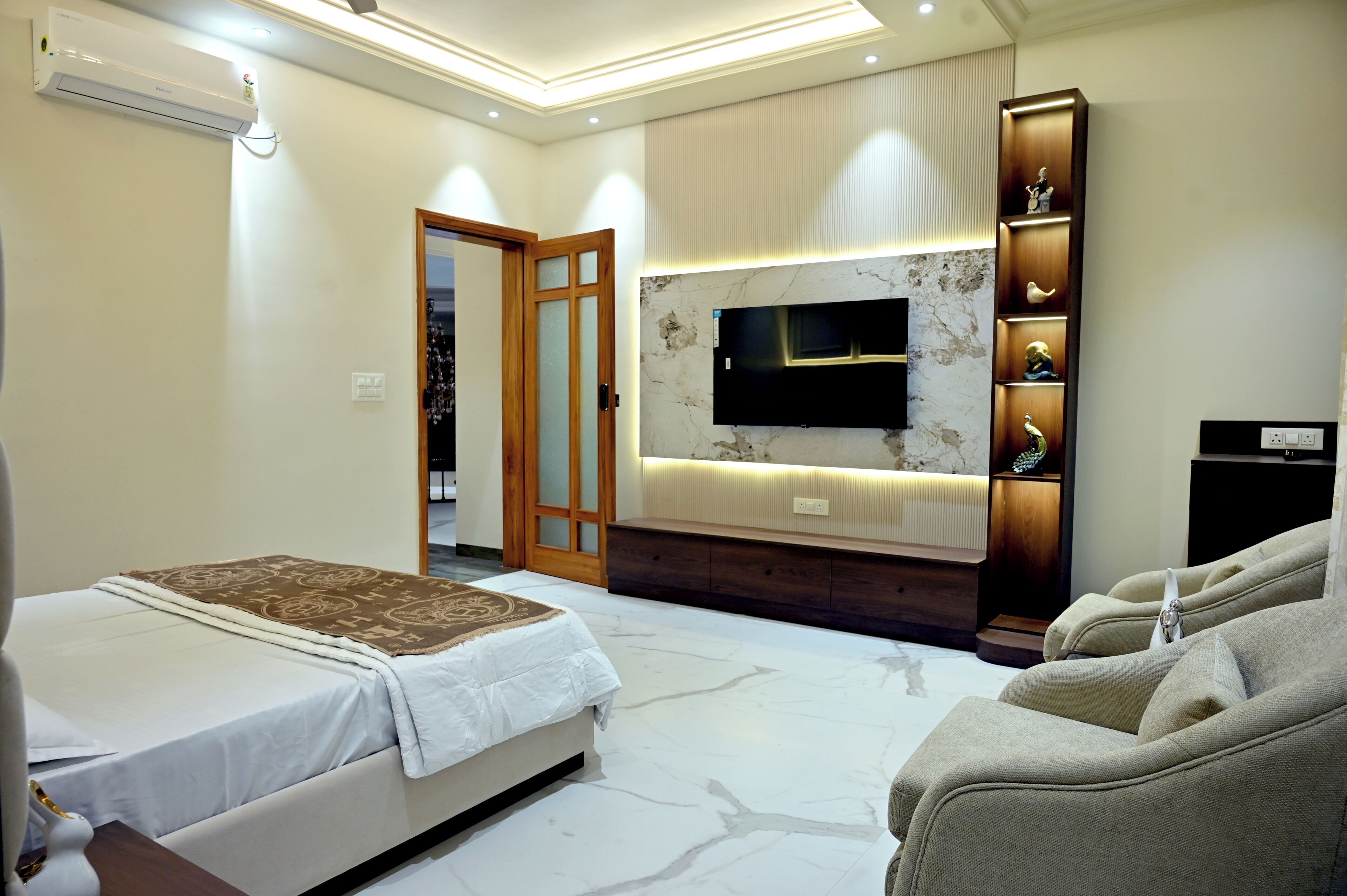 Un dormitorio moderno con cama, televisor montado en la pared, estantería empotrada con objetos decorativos y una puerta que da a otra habitación.