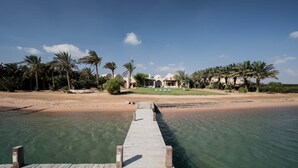 A wooden pier leading to a beach with clear water, palm trees, and a building with a white facade in the background. - Beit El Mona Sanctuary (Sea & Beach Front) (Hurghada)