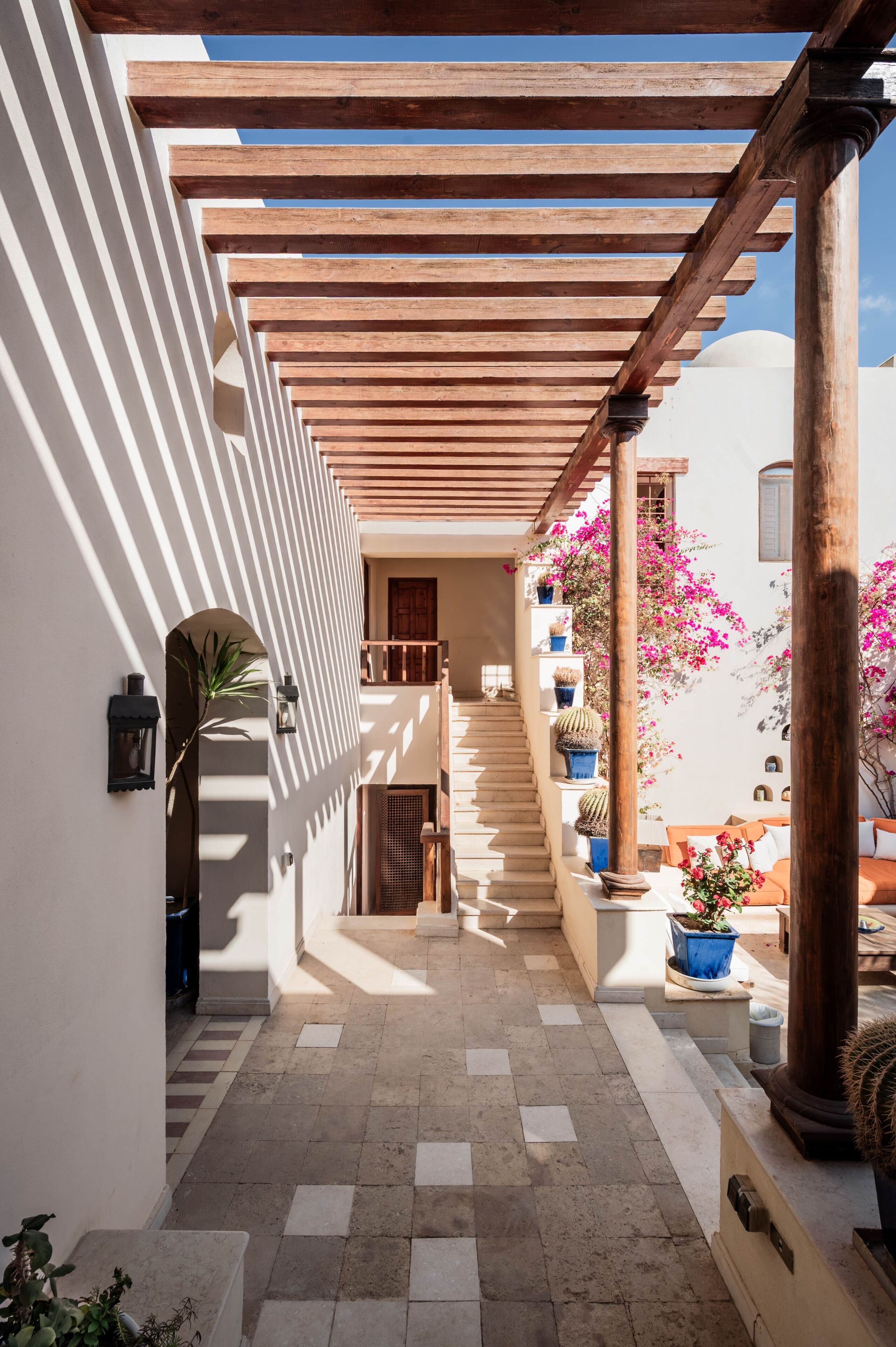 Un pasillo exterior cubierto con una pérgola de madera, suelo de baldosas y plantas en macetas.