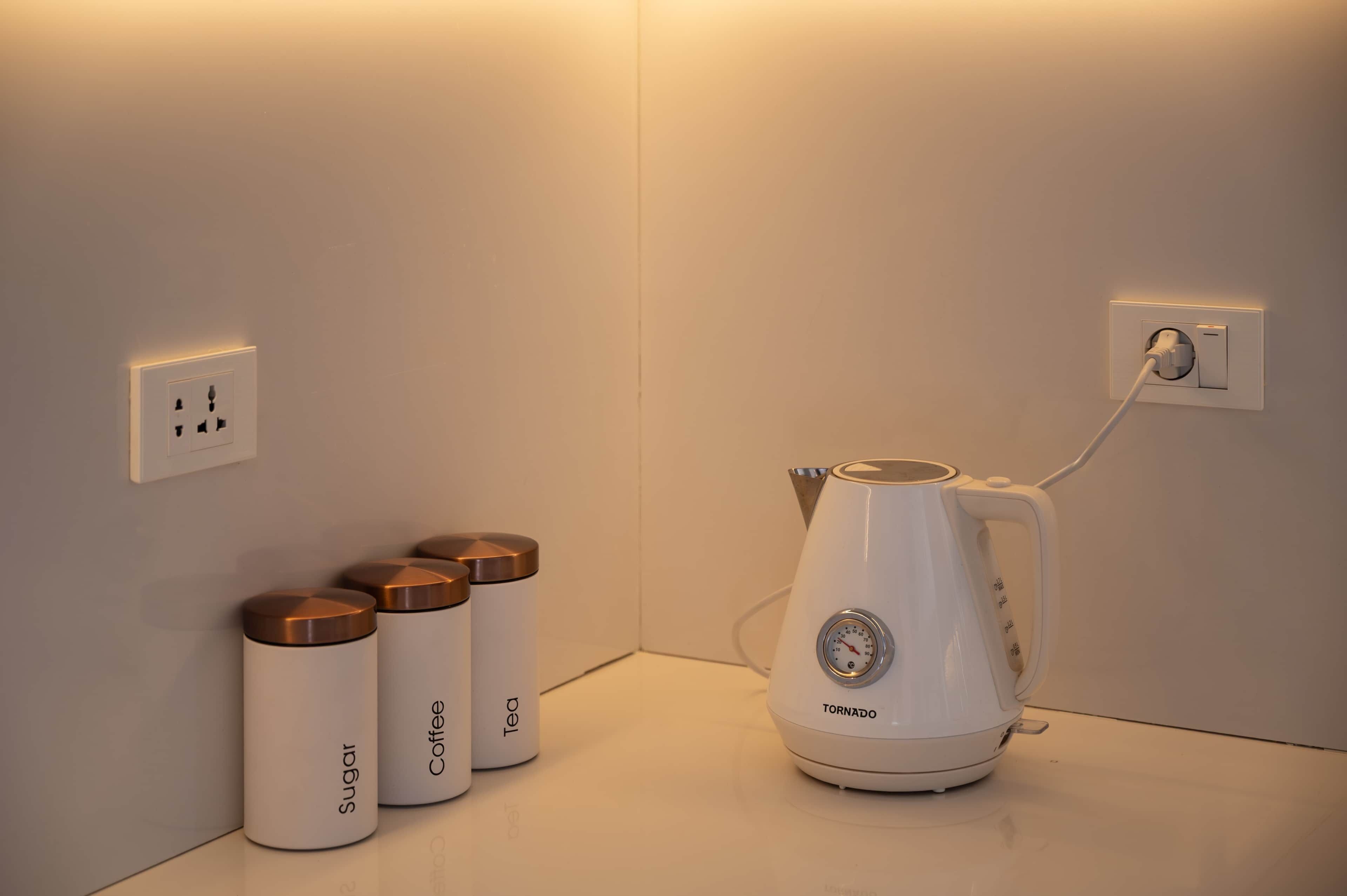 Une bouilloire blanche avec un cordon d’alimentation branché sur une prise murale, et trois récipients blancs avec des couvercles bruns, étiquetés « Sucre », « Café » et « Thé ».