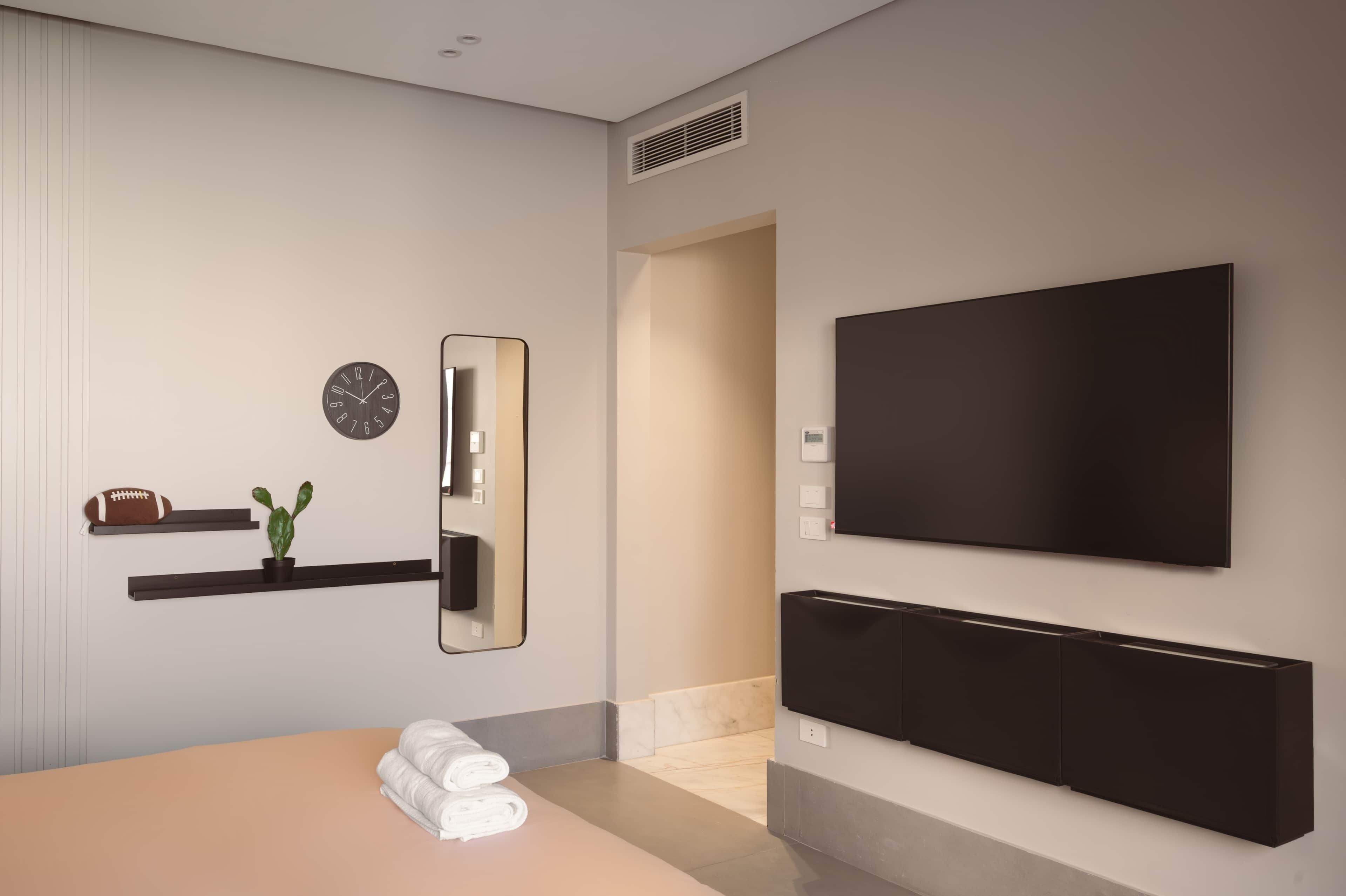 A modern hotel room with a large flat-screen TV, a clock, a mirror, a plant, and neatly folded towels on the bed.