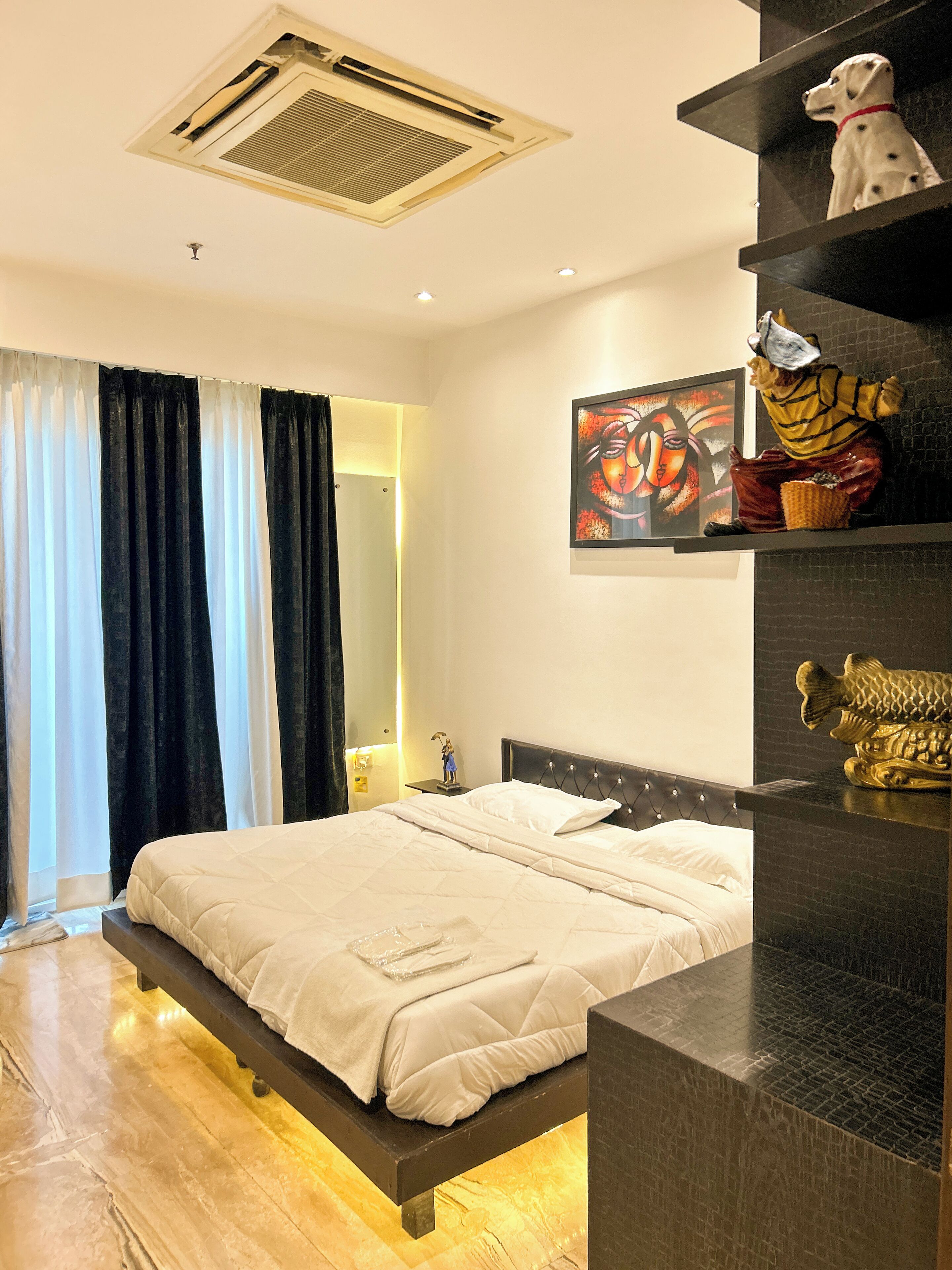 A modern hotel room with a large bed, a headboard with lights, a wall-mounted artwork, and a shelf with decorative items.