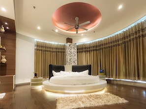 A modern bedroom with a circular bed, a ceiling fan, and a large window with curtains.