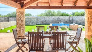 Un patio al aire libre con una mesa y sillas de madera, un muro de piedra y una piscina al fondo.