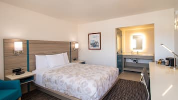 Habitación de hotel con cama, mesita de noche, lámpara y baño con espejo y lavamanos.