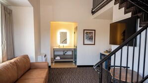 A hotel room with a brown leather sofa, a small bathroom with a mirror, a sink, and a framed picture on the wall. - Lemon Heights Inn (Tustin)