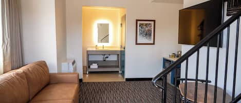 A hotel room with a brown leather sofa, a small bathroom with a mirror, a sink, and a framed picture on the wall.