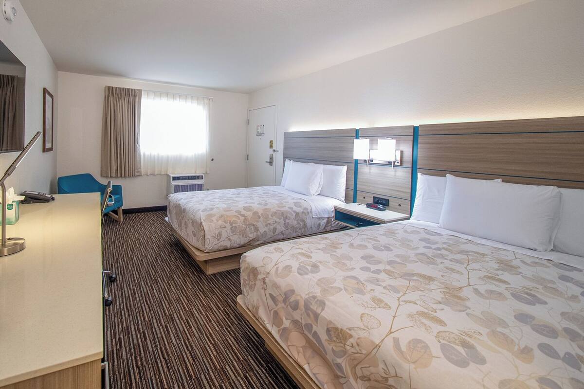 Une chambre d’hôtel avec deux lits, un bureau, une chaise, une fenêtre avec des rideaux, un climatiseur et une lampe fixée au mur.