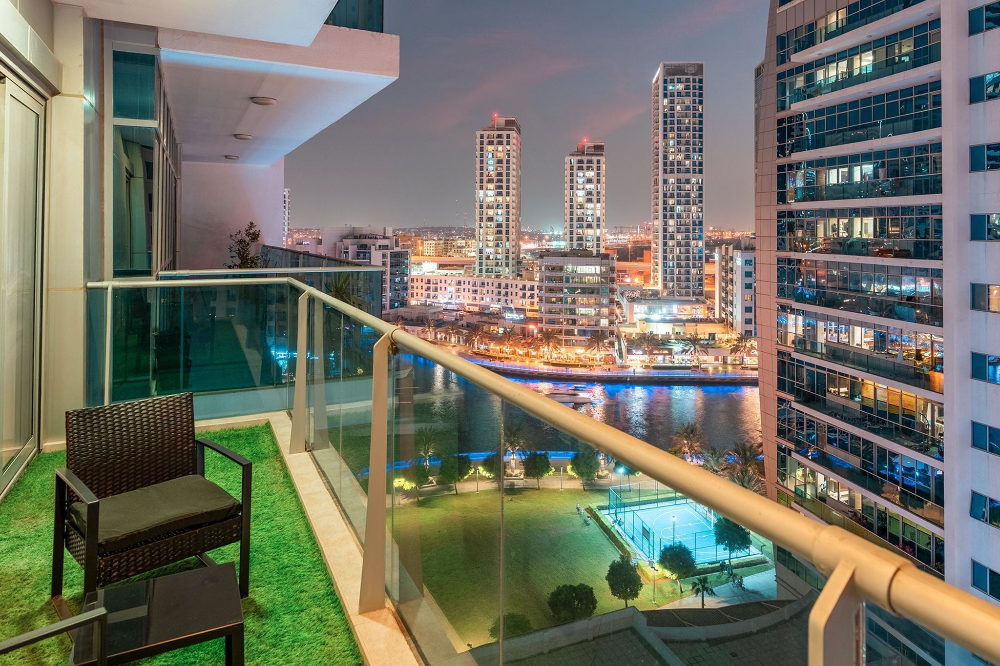 Een balkon met glazen hekwerk met uitzicht op een stadsbeeld bij nacht, met moderne gebouwen en een rivier.