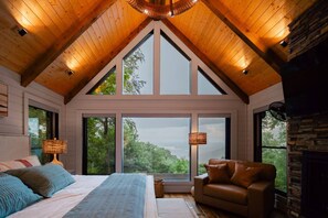 A bedroom with a sloped ceiling, large windows, a bed, a chair, and a lamp. - Wellness Retreat: Epic Views·Gym·Sauna·Cold Plunge (Blue Ridge)
