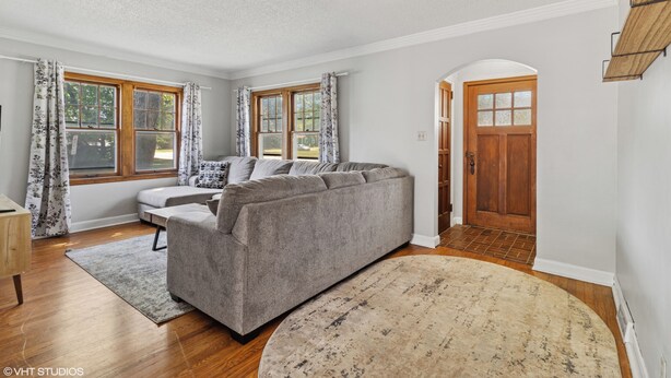 A modern living room with a grey sectional sofa, a small white table, and a large round rug. There are two windows with floral curtains, a wooden door, and a white ceiling. - Beautiful 3-bedroom house with AC in adorable Des Moines (Des Moines)