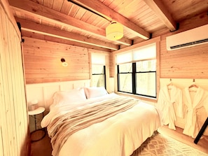A wooden cabin bedroom with a bed, a bedside table, a hanging robe rack, and a window with a view of trees.