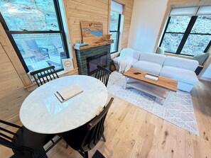 Ein gemütliches Wohnzimmer mit Kamin, einer Couch, einem runden Tisch und Blick ins Grüne durch große Fenster.