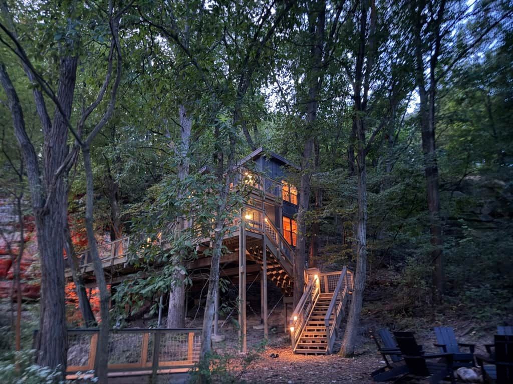 Una casa en árbol con escalera de madera y terraza, rodeada de un bosque denso.