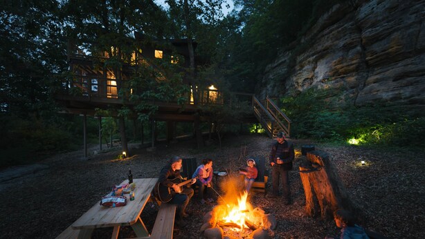 Eine rustikale Outdoor-Umgebung mit einem Holztisch, einem Lagerfeuer und einer Holzterrasse mit einer Treppe zu einem Haus.