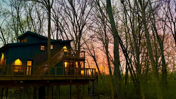 Une cabane en bois dotée d’un balcon, entourée d’arbres, au coucher du soleil.