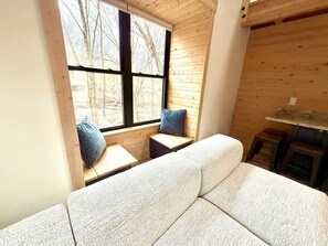 Een kamer met een bank, twee ramen met zonwering, een bankje met kussens en een klein krukje.
