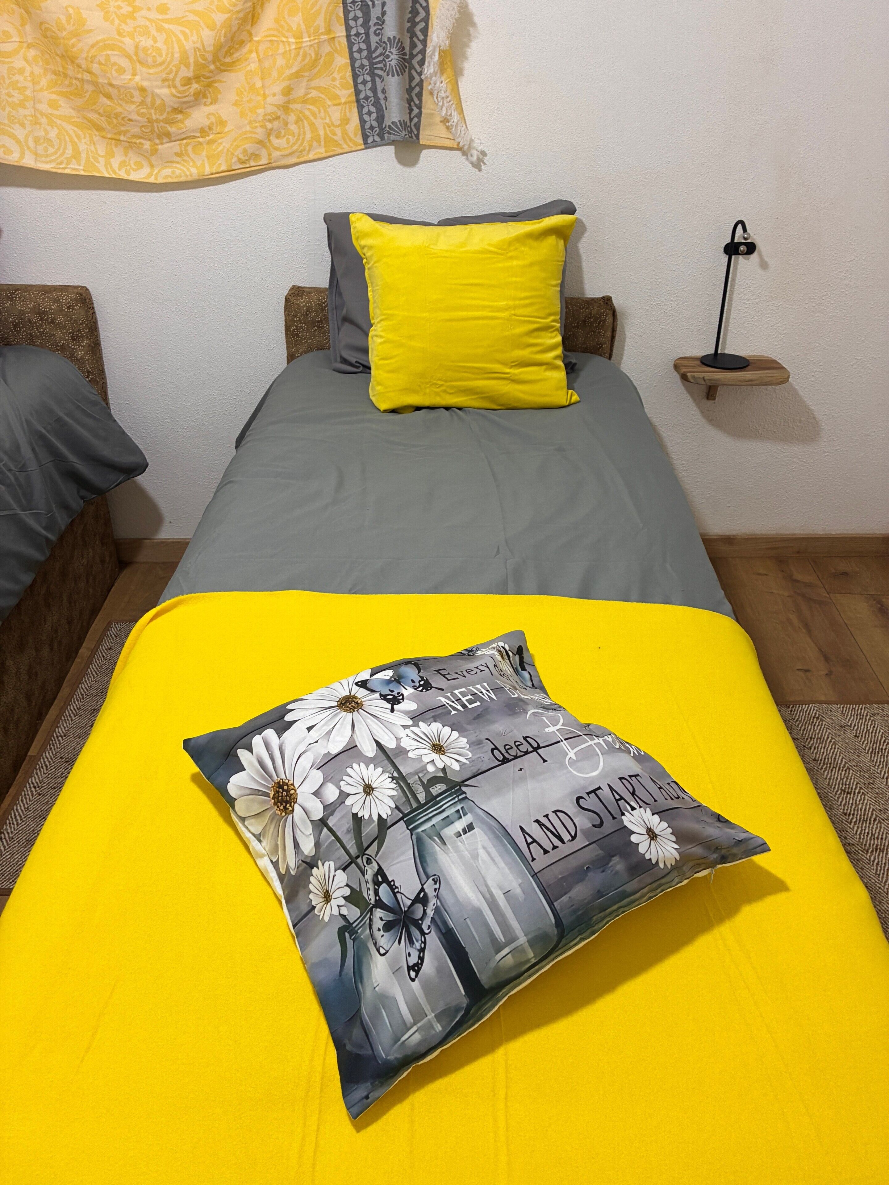Una cama con cubrecama amarillo, un cojín con diseño floral y de botellas, y una mesita de noche con una lámpara.