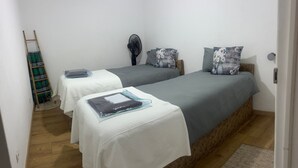 Ein Zimmer mit zwei Betten, jedes mit einer weißen und grauen Tagesdecke, einem Nachttisch mit einem Buch und einem Wandventilator.
