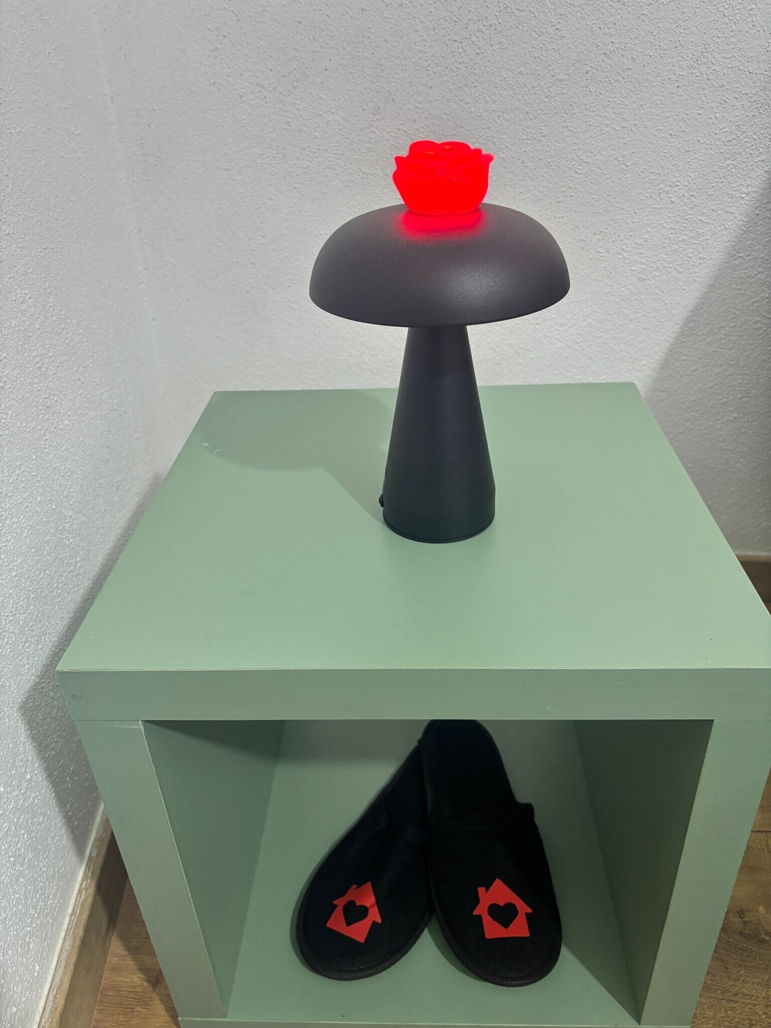 Een groene nachtkastje met een zwarte lamp en een paar zwarte pantoffels versierd met rode hartjes.
