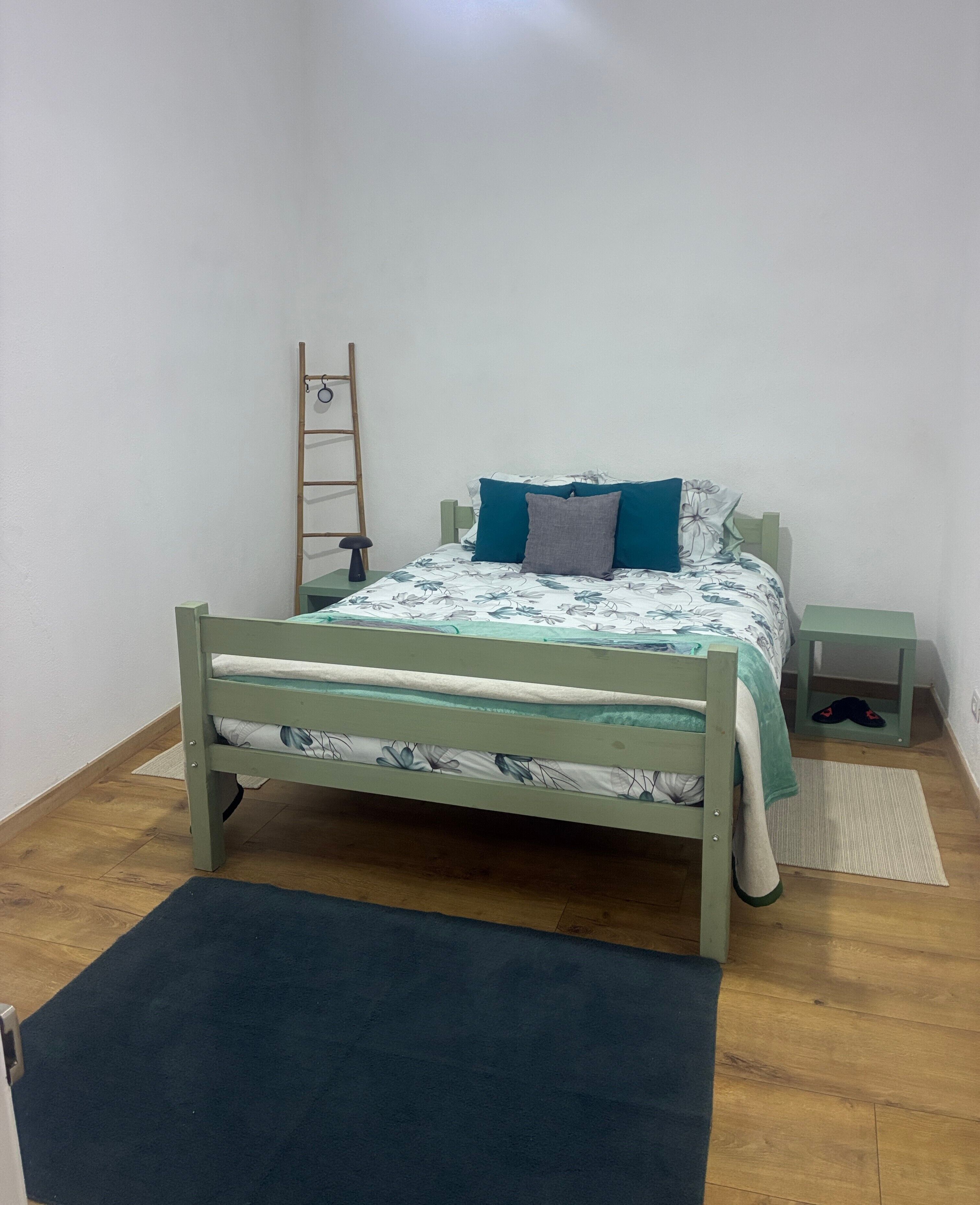 Een enkel bed met een bloemenprint deken, een houten ladder, een klein nachtkastje en een donker vloerkleed op een houten vloer.