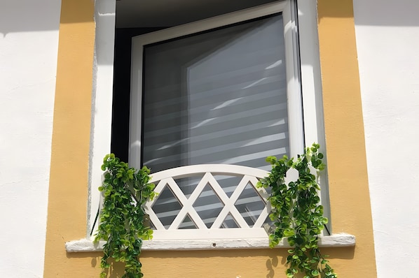 Una ventana con marco blanco y una reja de diseño de rombos, flanqueada por plantas verdes.