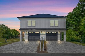 A modern, two-story duplex is pictured at dusk, with a colorful pink and purple sky above. The building has a dark metal roof and light gray siding. Each unit features a two-car garage with a separate paved driveway. - Towne Treasures A ~ Brand-New Luxe Coastal Retreat with Private Cocktail Pool (Panama City Beach)