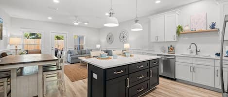 An open-concept living area features a kitchen with a large dark gray island and white cabinets. The space extends into a dining area and living room with a gray sectional sofa, all on light wood-look flooring.