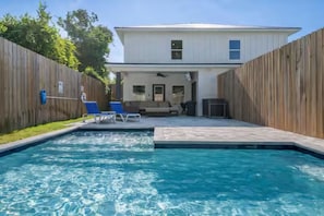 A bright backyard features a small pool with a paver deck. A row of blue Adirondack chairs sits by the water, with two white and blue lounge chairs in the foreground. A tall wooden fence encloses the yard, and a large tree is visible beyond the fence. - Towne Treasures A ~ Brand-New Luxe Coastal Retreat with Private Cocktail Pool (Panama City Beach)
