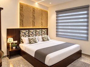 A hotel room with a large bed, a nightstand with a lamp, a wall-mounted artwork, and a window with blinds. - Hotel Destin (Gwalior)