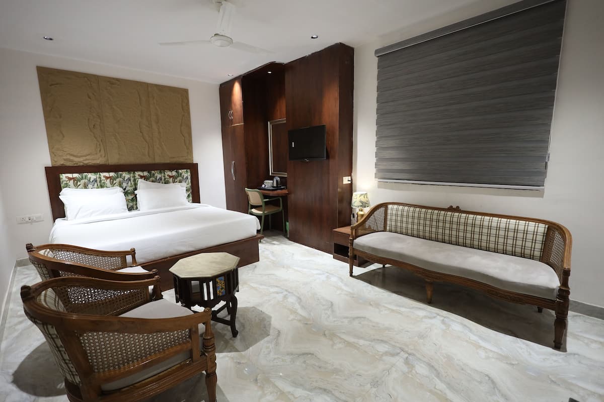 A bedroom with a bed, two wooden chairs, a small table, a sofa, a television, and a large window with blinds.