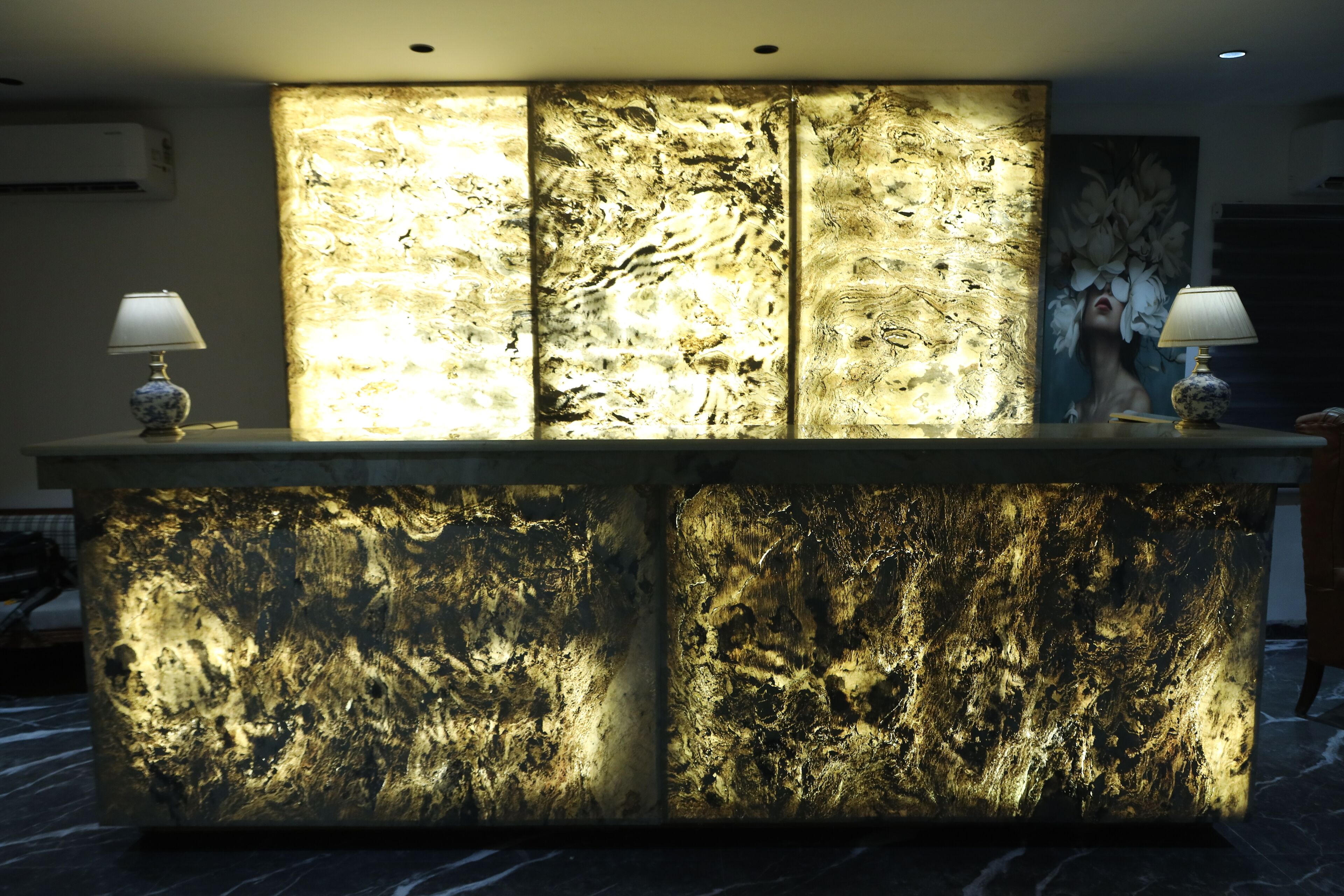 A reception desk with a marble-like backlit panel, two lamps, and a large floral painting on the wall.