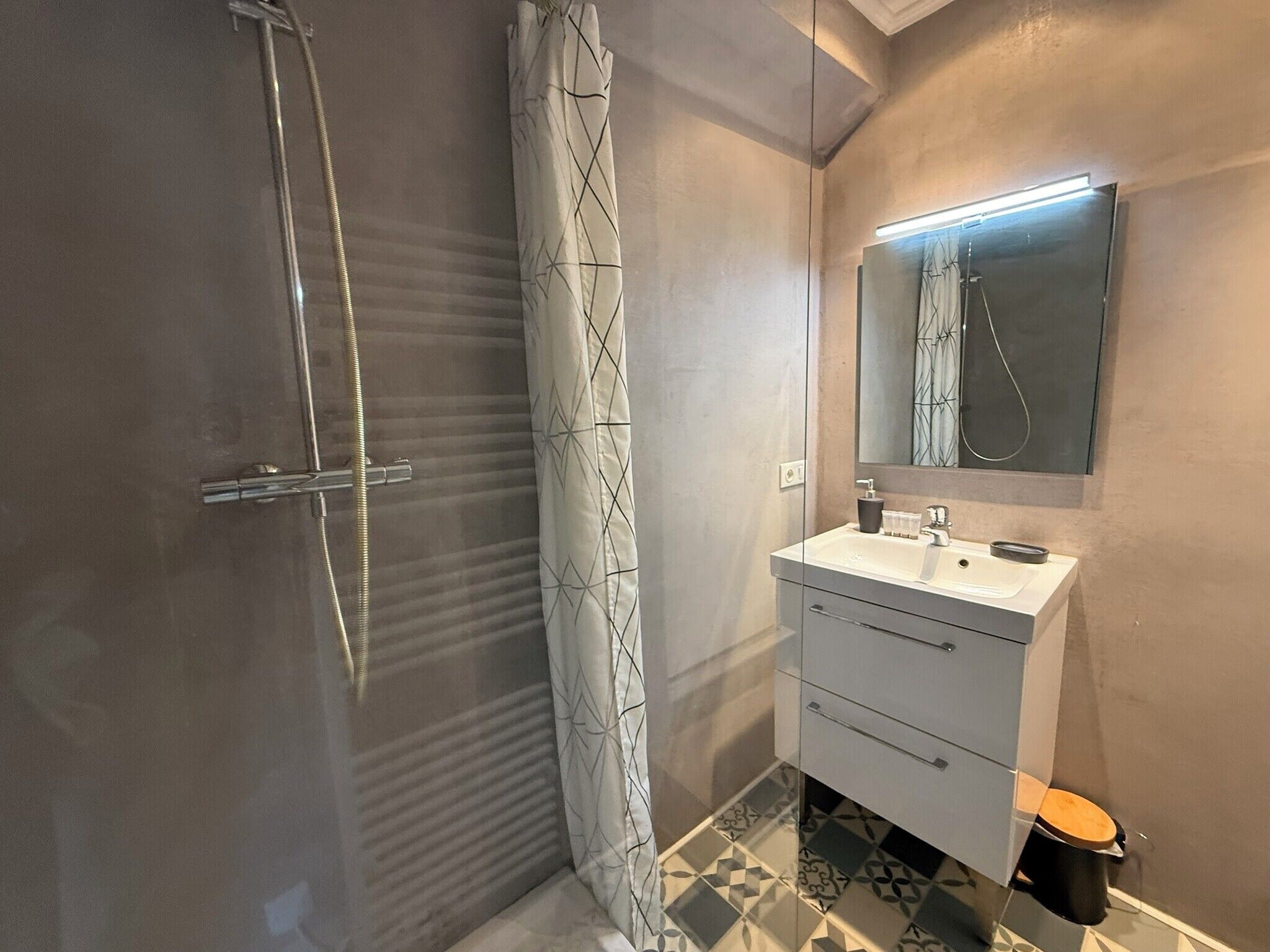 Een moderne badkamer met een glazen douchecabine, een badgordijn met een patroon, een wit wastafelmeubel met een spiegel en een toilet met een houten zitting.