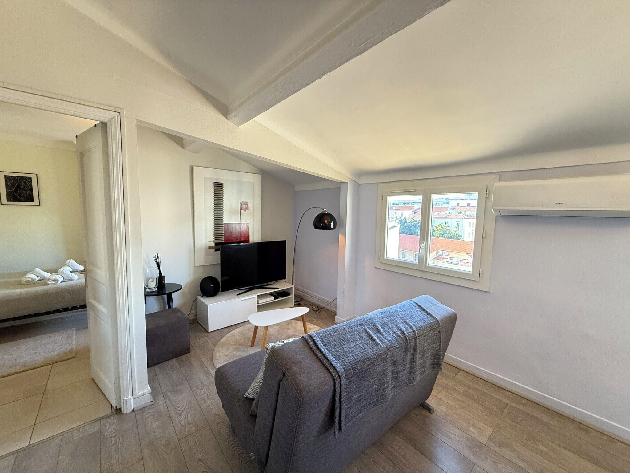 A modern living room with a grey sofa, a flat-screen TV, a small white table, and a floor lamp. There is a window with a view of buildings outside.