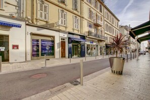 A street with buildings, a large metal planter with a tree, and a blue awning over a shop. - Nice one-bedroom apartment near the Palais des Festivals (Cannes)