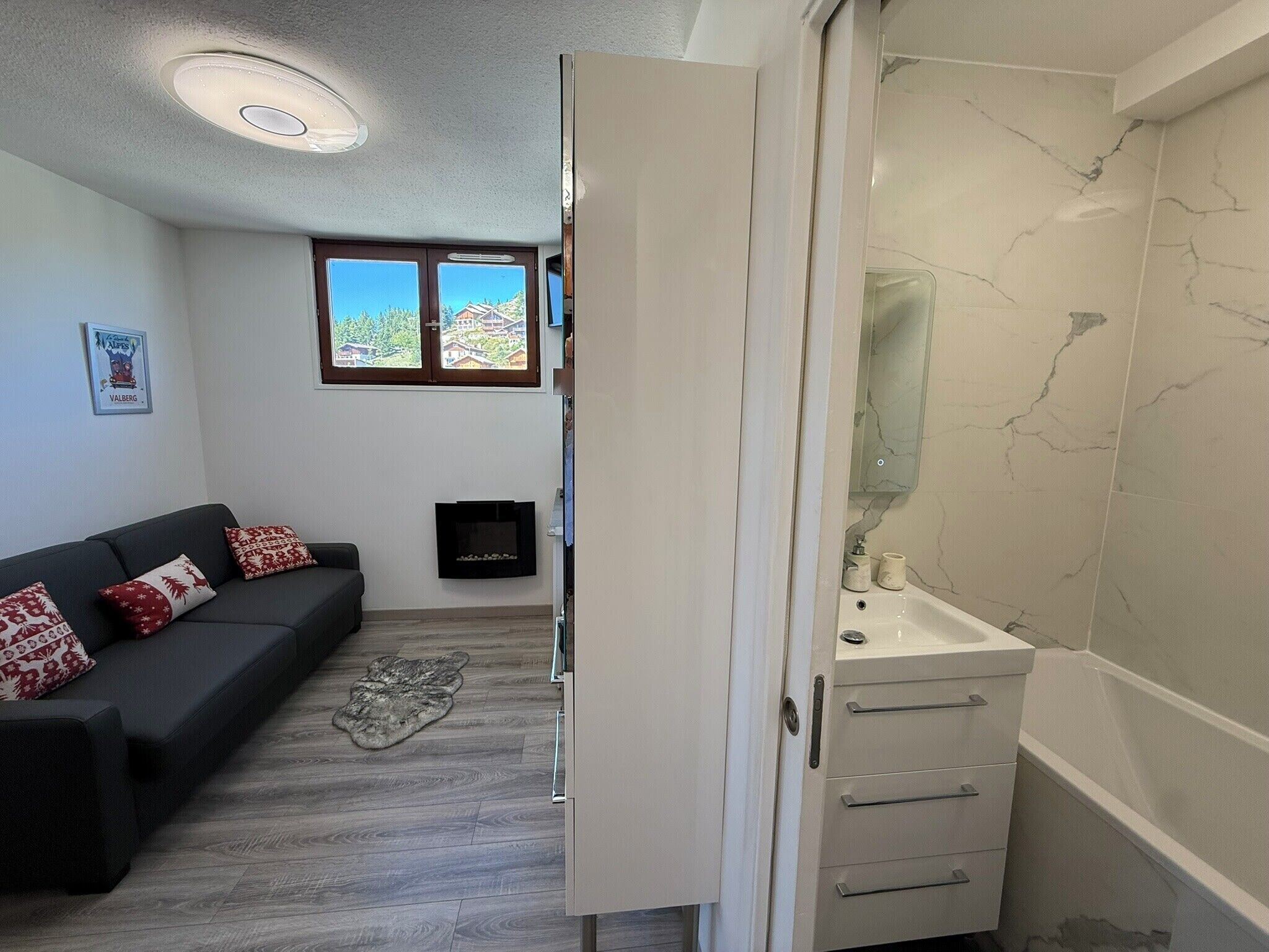 A modern room with a grey sofa, a small rug, a fireplace, and a bathroom with a marble wall and sink.